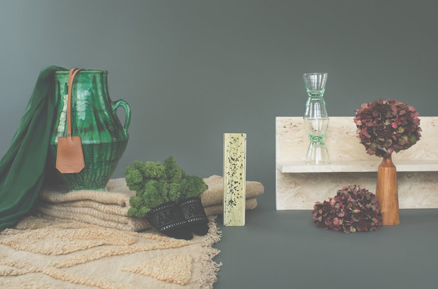 Still life on a slate-grey backdrop: a green glazed jug with a leather tag, folded wool blankets, moss, glassware, a travertine shelf and dried hydrangeas