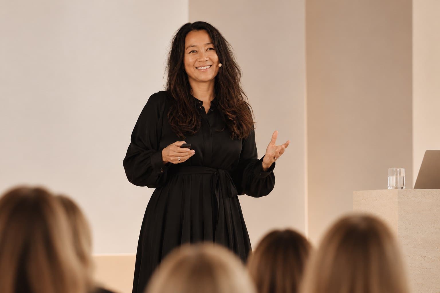 Anne Marie Mondrup smiling as she speaks to an audience, in a black dress with a headset microphone and a presentation clicker